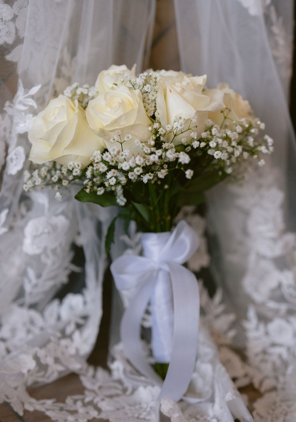 White rose bridal bouquet with baby's breath, tied with a white satin ribbon, against a lace wedding dress backdrop.