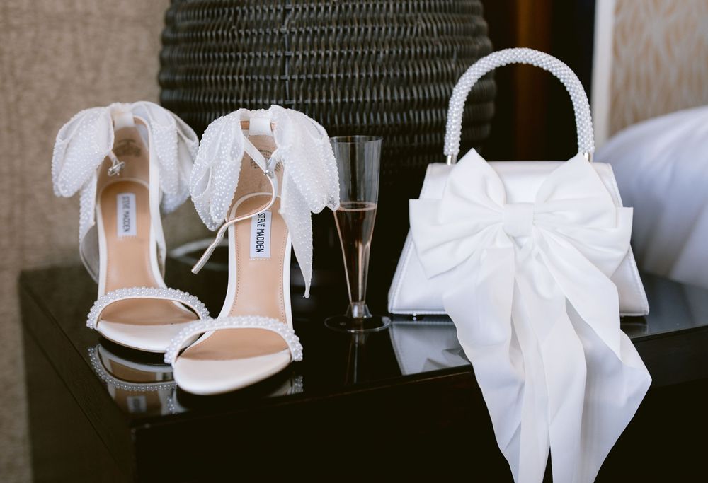 White bridal heels with feather details and white bow clutch purse on black table with champagne glass.