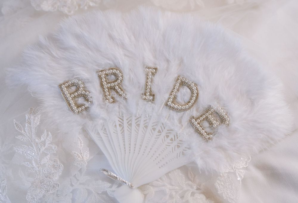 White feather bridal fan with pearl and rhinestone BRIDE letters, elegant wedding accessory on lace background.