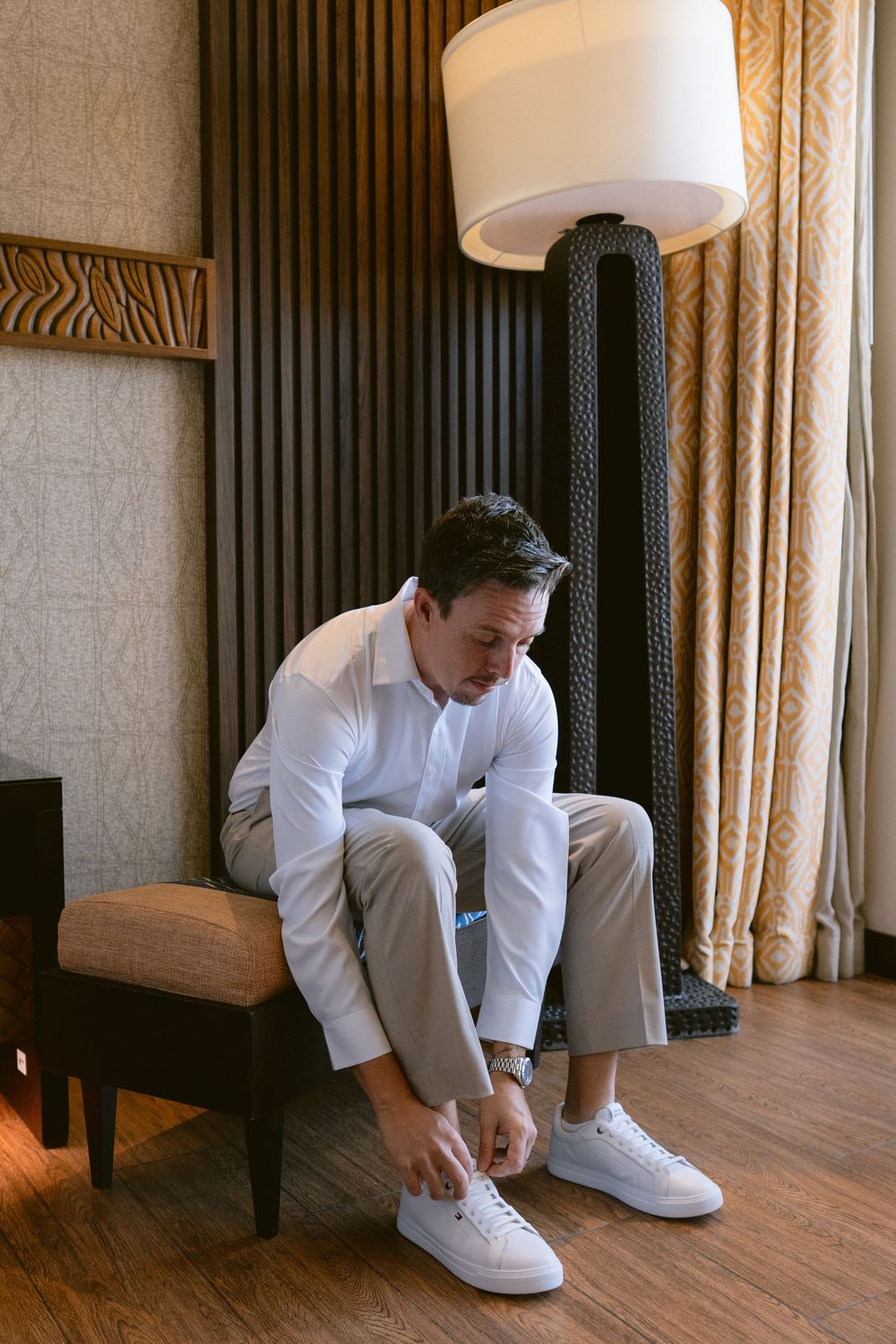 Man in white shirt and grey trousers tying white sneakers while seated in an elegant hotel room.