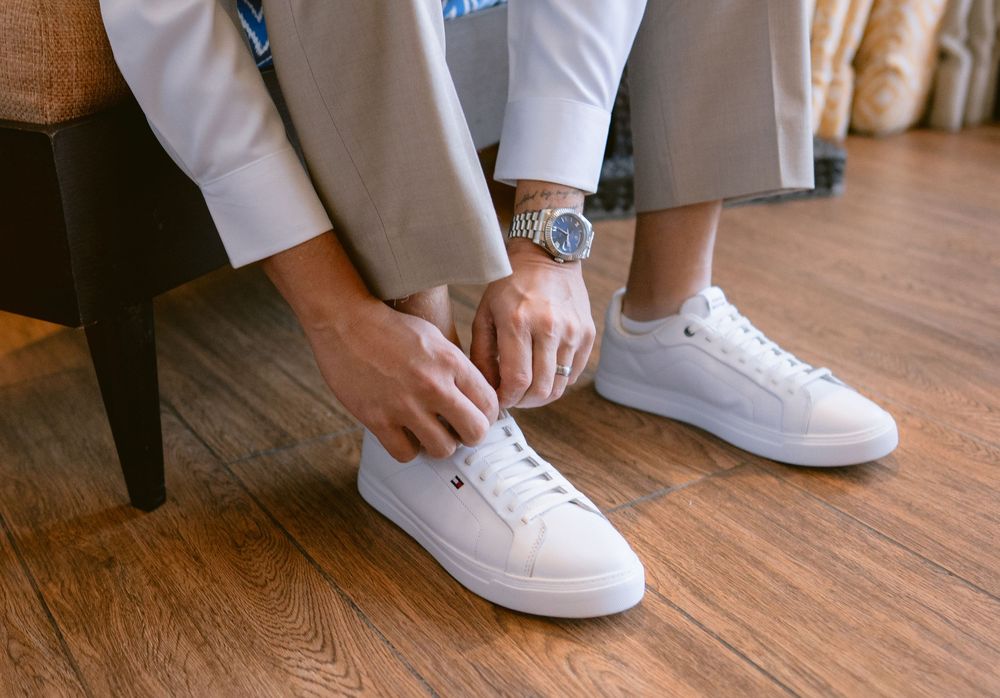 Man in gray suit tying white leather sneakers on hardwood floor, wearing a silver watch.