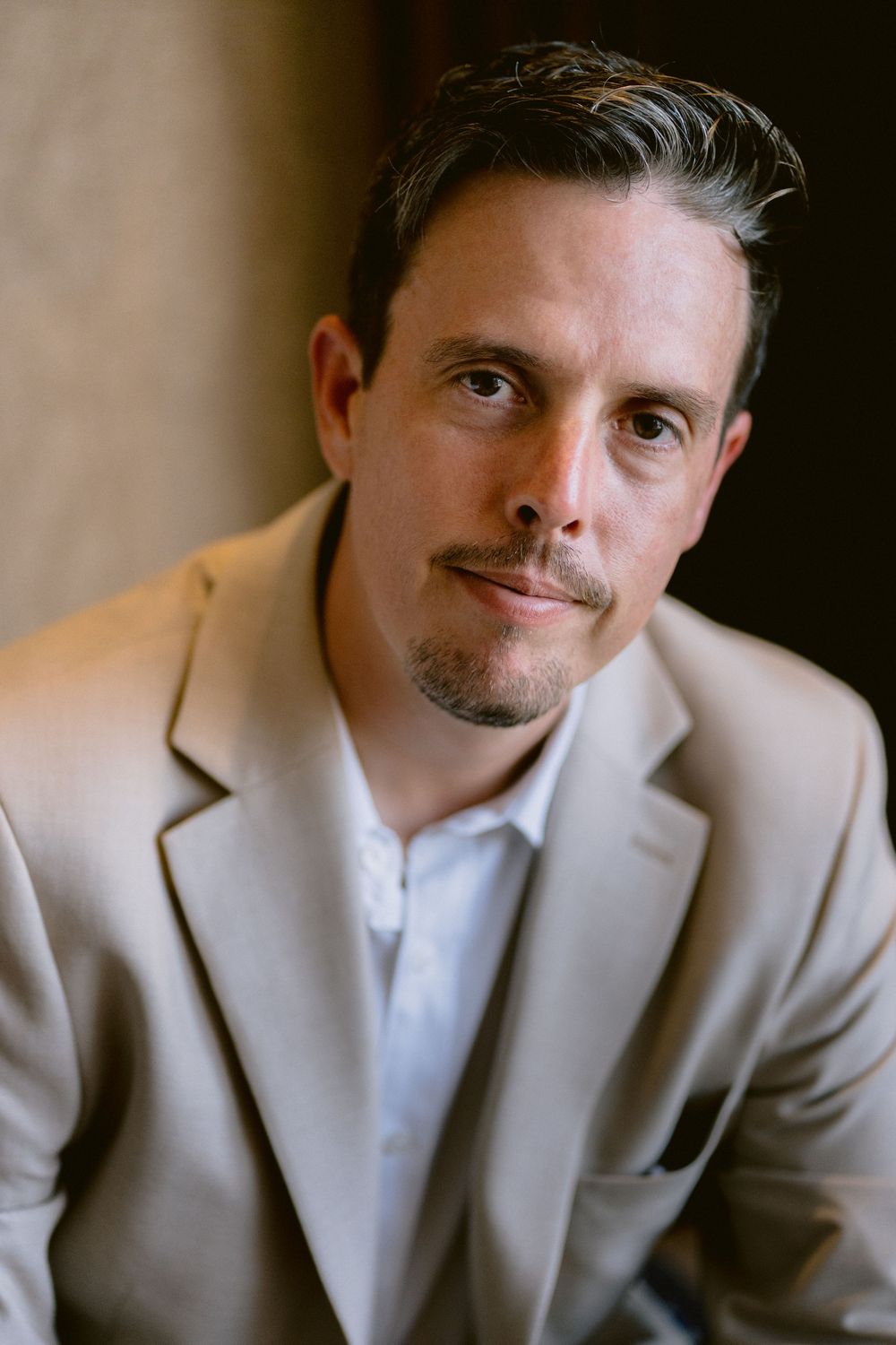 Professional headshot of a man in a beige blazer with dark hair and goatee, looking confidently at the camera.