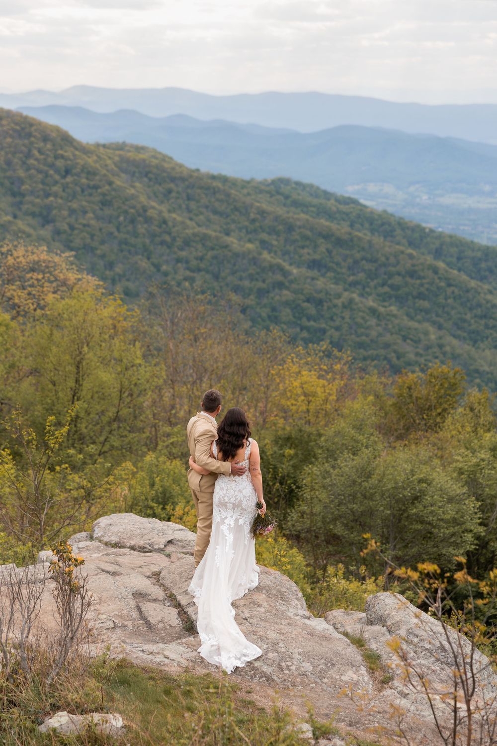 Bride and groom embracing on rocky mountain overlook with lush green valleys and blue ridges in background during their Shenandoah wedding.