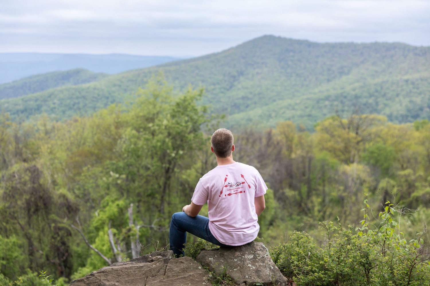 High school graduate sitting on rock overlooking lush green mountain landscape on a cloudy day at an overlook in Shenandoah National Park.