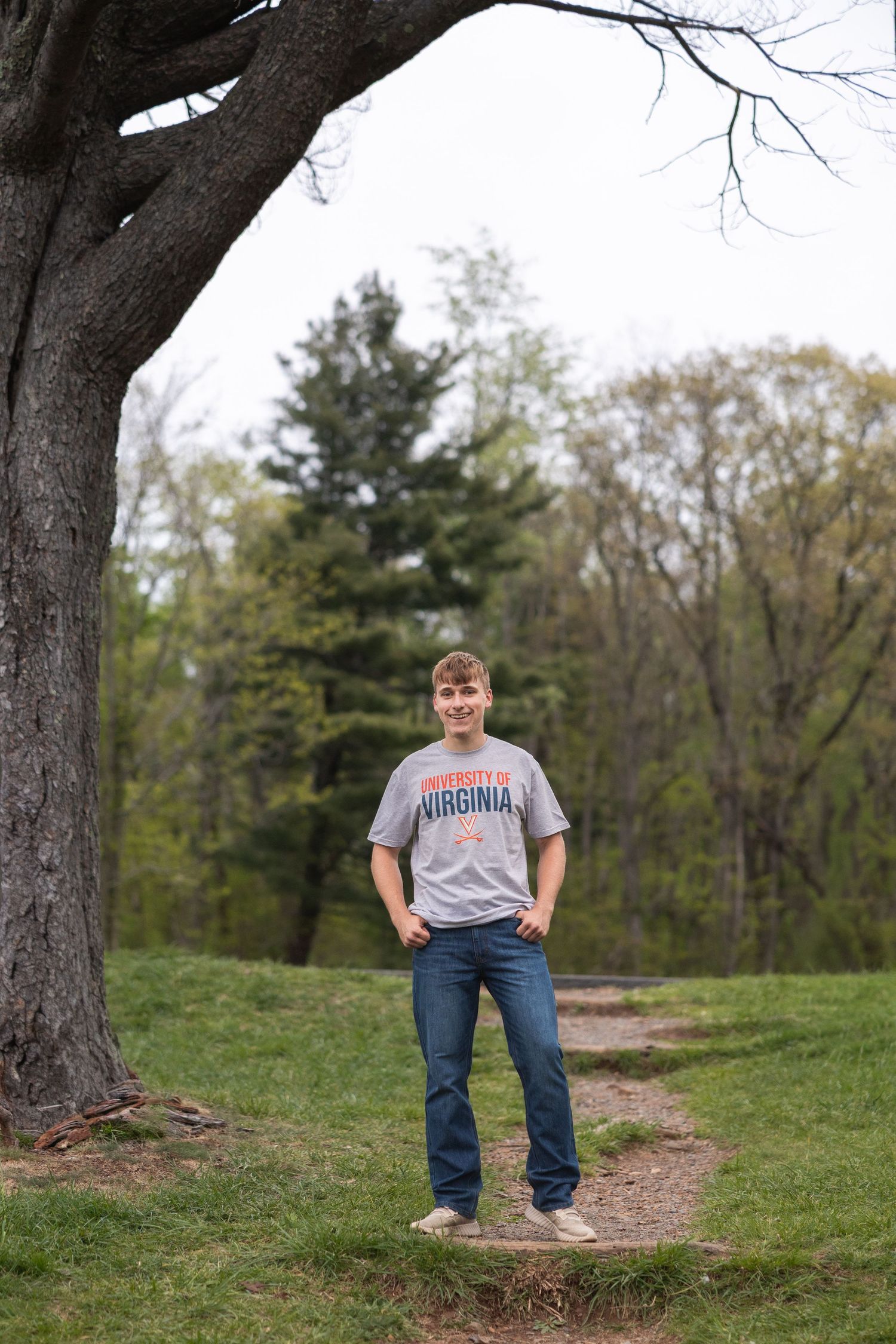 Young man in University of Virginia t-shirt and jeans standing on a path beside a large tree in a green park.