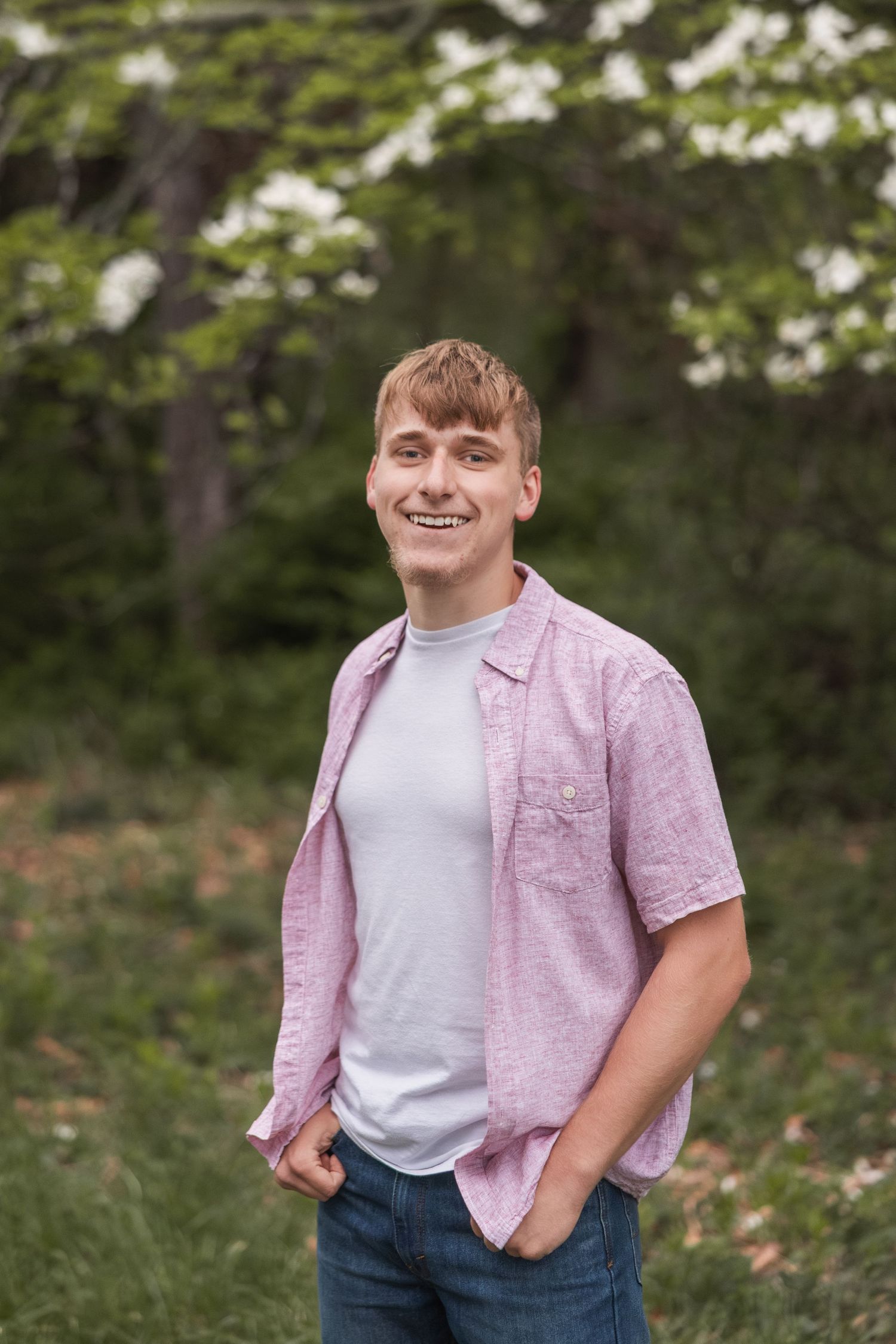 Smiling young man in pink shirt and jeans standing outdoors among green trees and white blossoms.