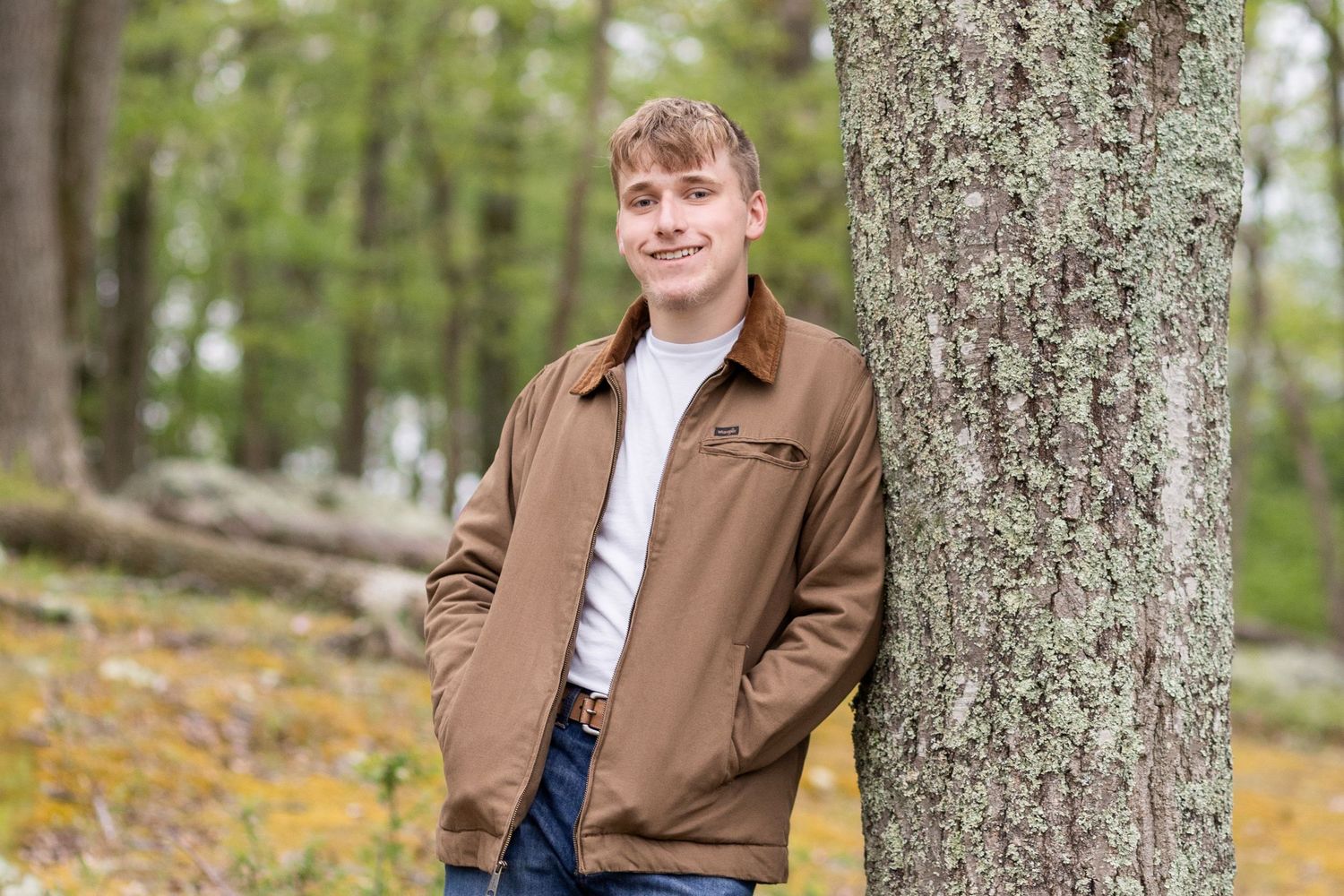 Young man in brown jacket leaning against a tree in a forest setting for a senior portrait photo.
