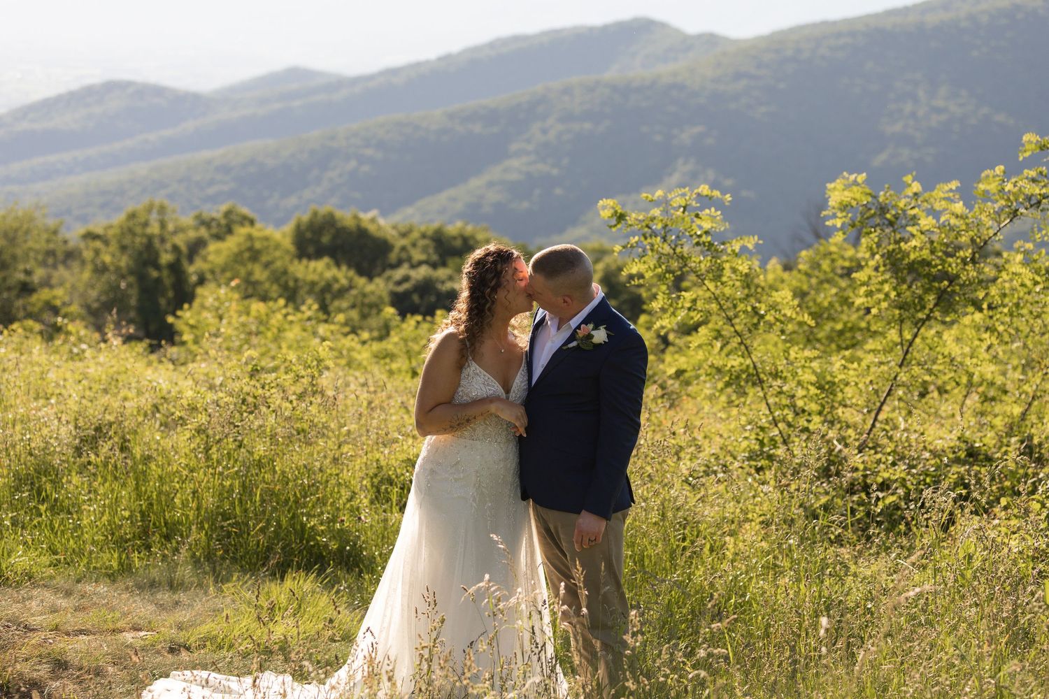 Bride and groom share a romantic kiss in a lush green meadow at Pass Mountain Overlook with scenic mountain views on their wedding day.