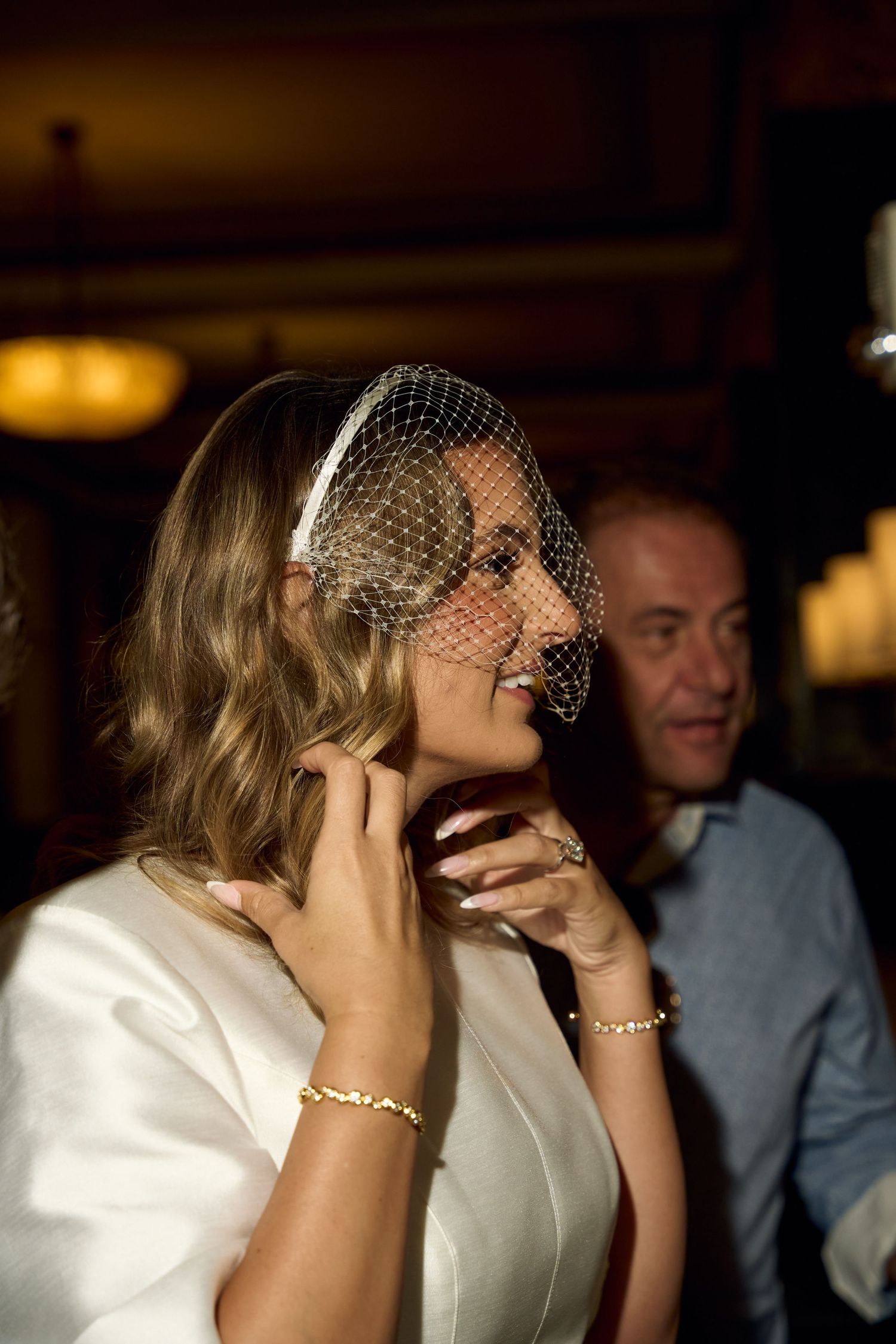 Smiling bride in white dress and birdcage veil adjusting her hair at an elegant wedding reception.