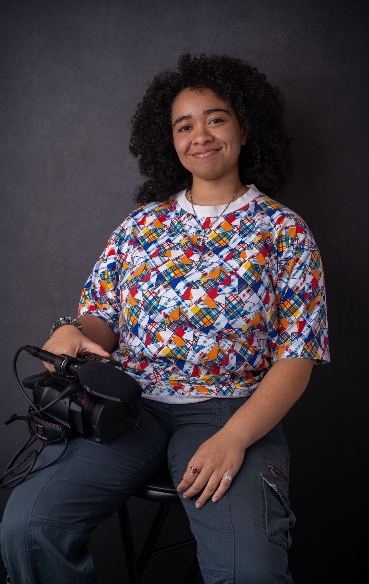Young woman with curly hair holding a camera, wearing a colorful geometric print shirt, seated against a dark background.