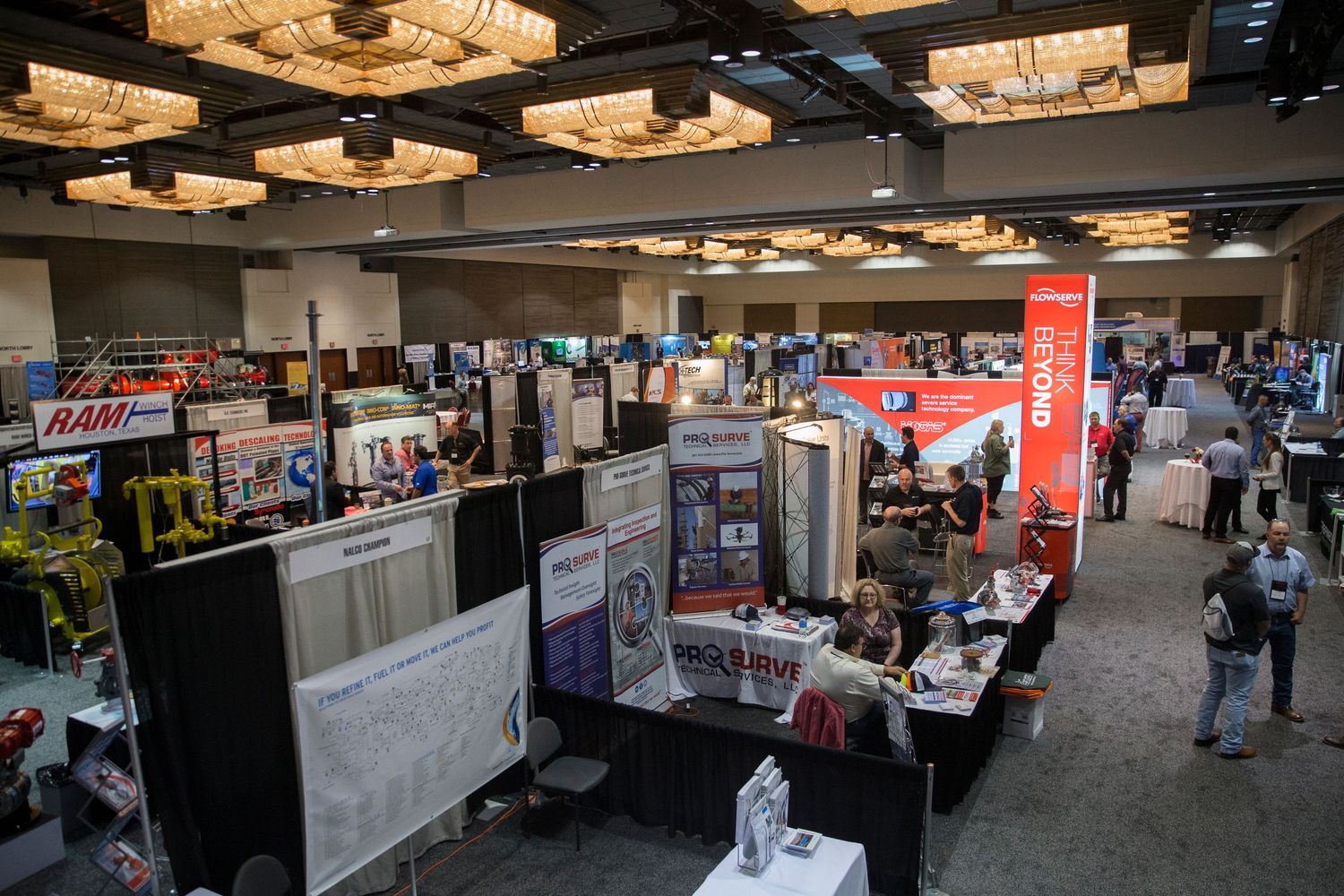 Busy trade show exhibition hall with vendor booths, signage, and attendees browsing displays under ornate ceiling lights.