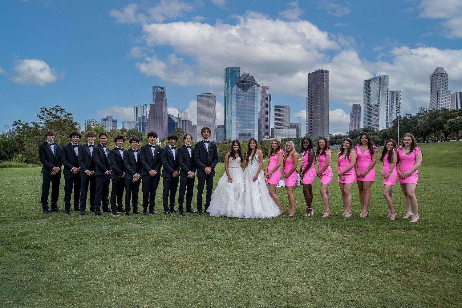 Quinceañera court poses with city skyline backdrop, boys in suits and girls in pink dresses on green lawn.