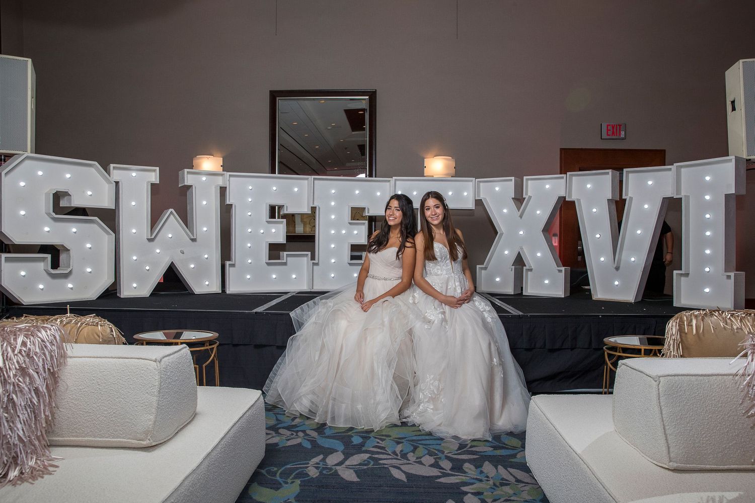 Two girls in white gowns pose in front of illuminated Sweet XVI marquee letters at a Sweet 16 party.