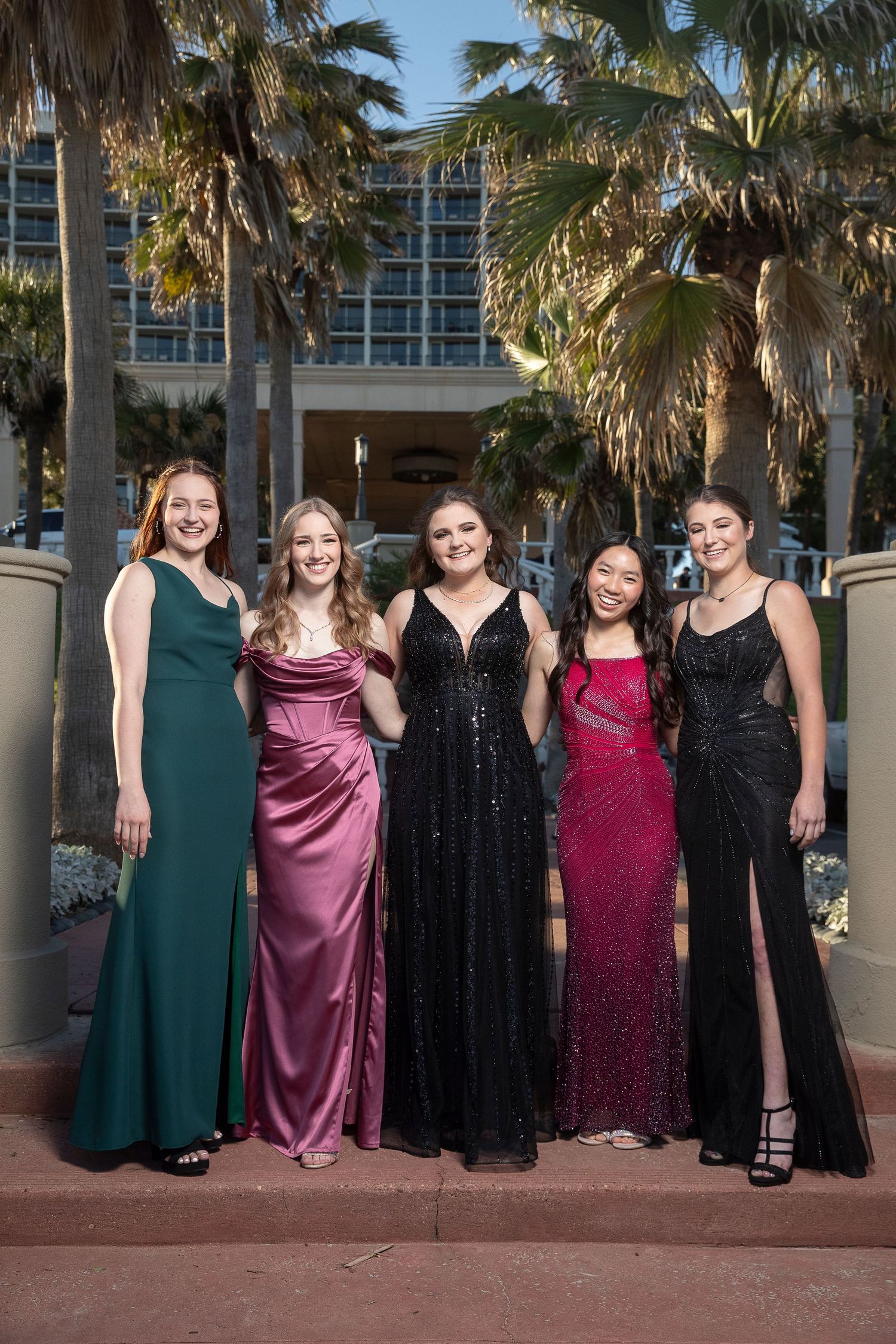 Five young women in elegant formal gowns pose together outdoors near palm trees at prom.