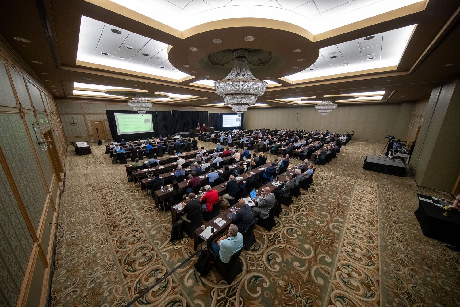 Large conference room filled with attendees seated at tables facing a presentation screen in an elegant ballroom.
