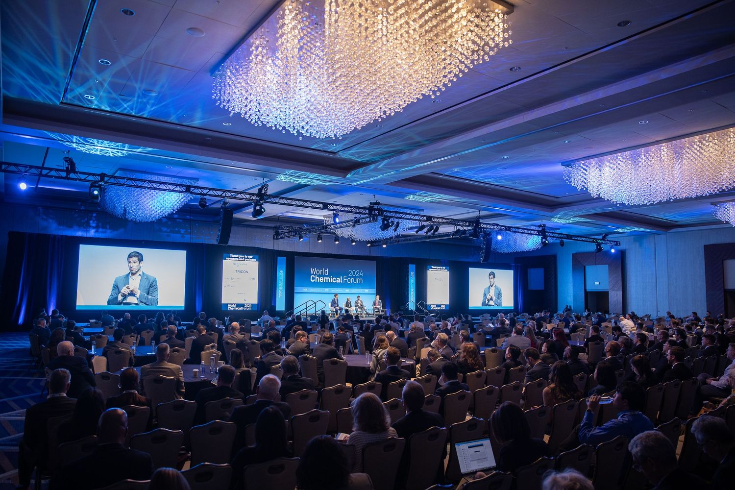 Large corporate conference in a ballroom with crystal chandeliers, audience seated before illuminated presentation screens.