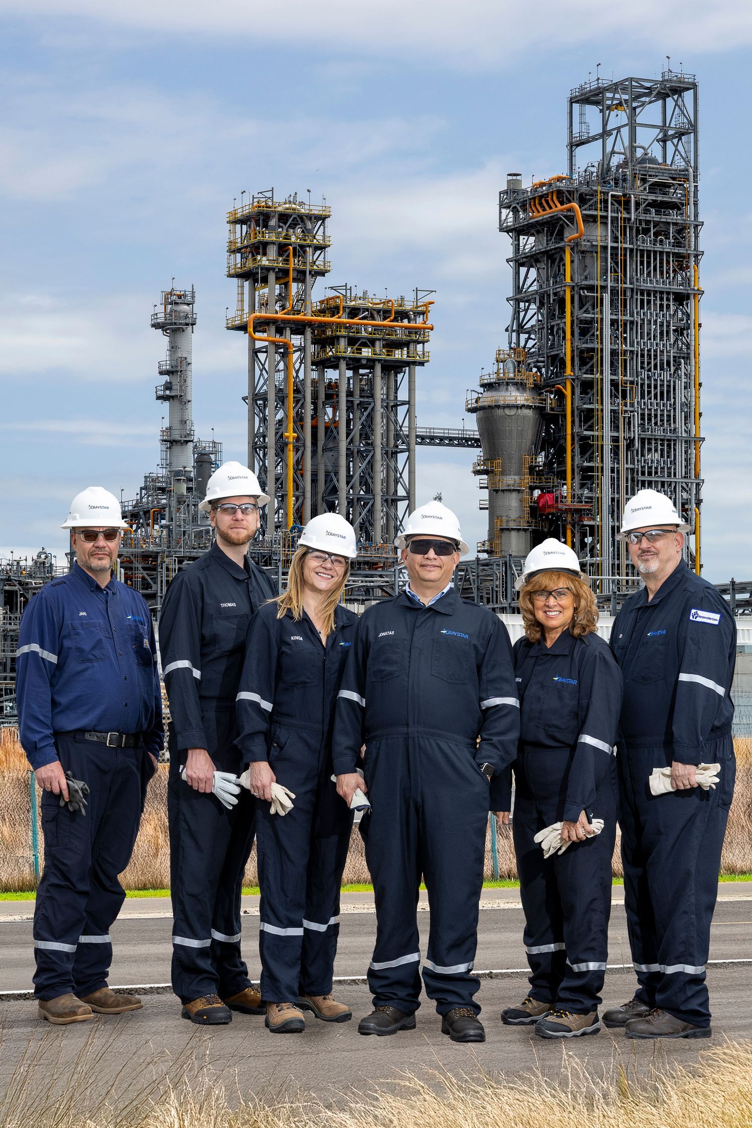 Six industrial workers in navy coveralls and white hard hats pose together in front of an oil refinery.