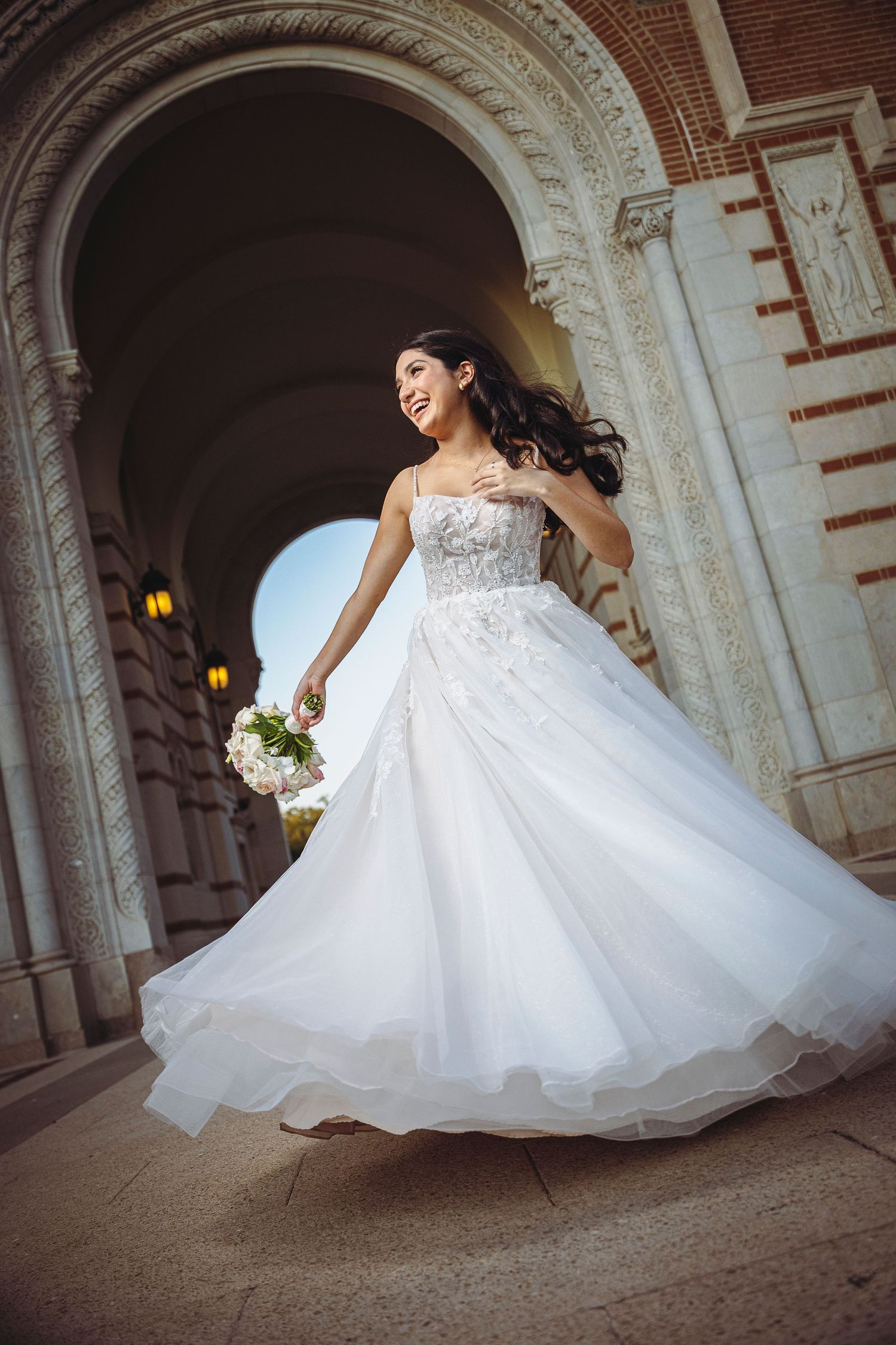 Bride in flowing white ball gown twirling under ornate stone arch, holding bouquet, smiling joyfully.