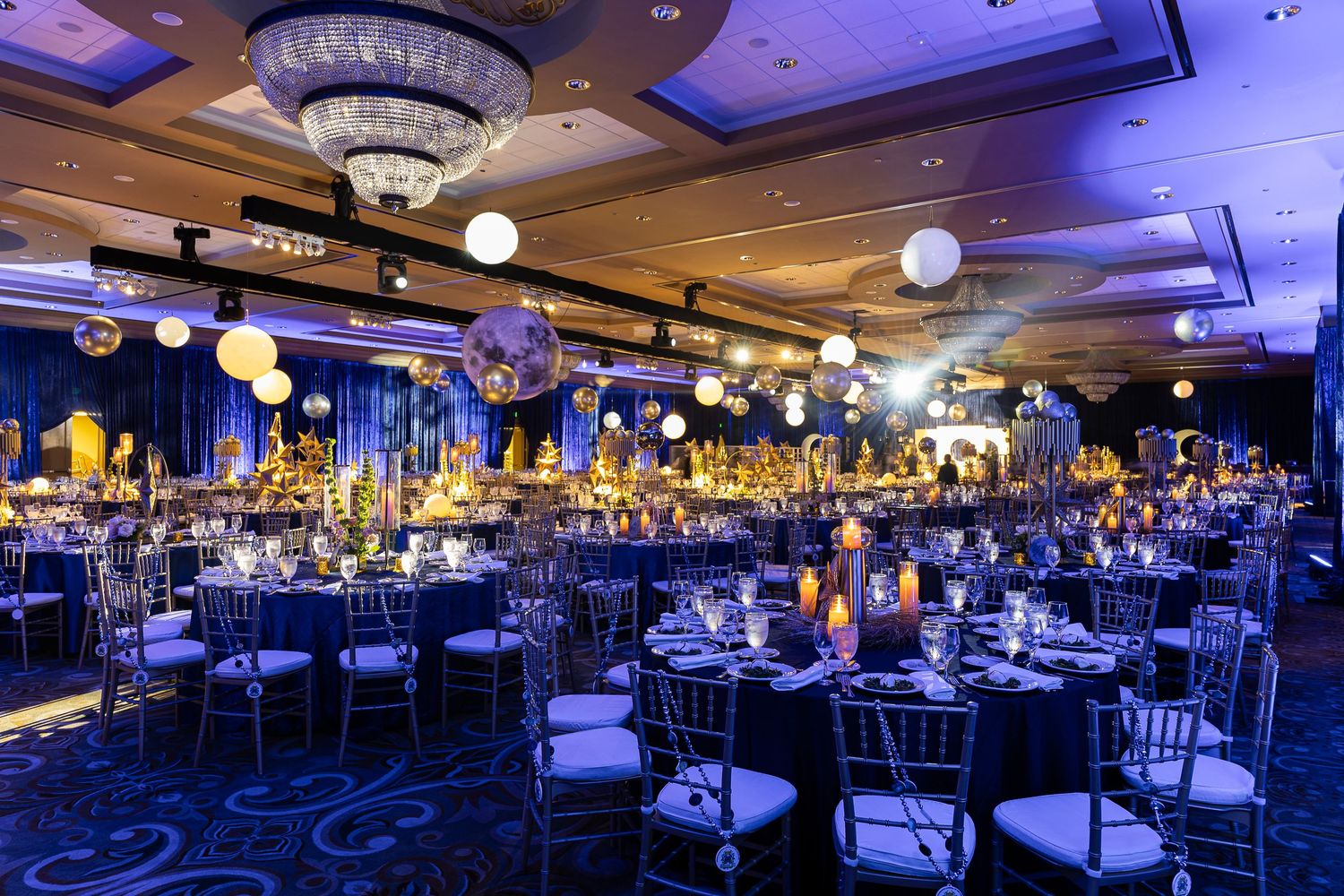 Elegant ballroom event setup with round tables, chiavari chairs, glowing lanterns, and blue lighting for a formal gala.