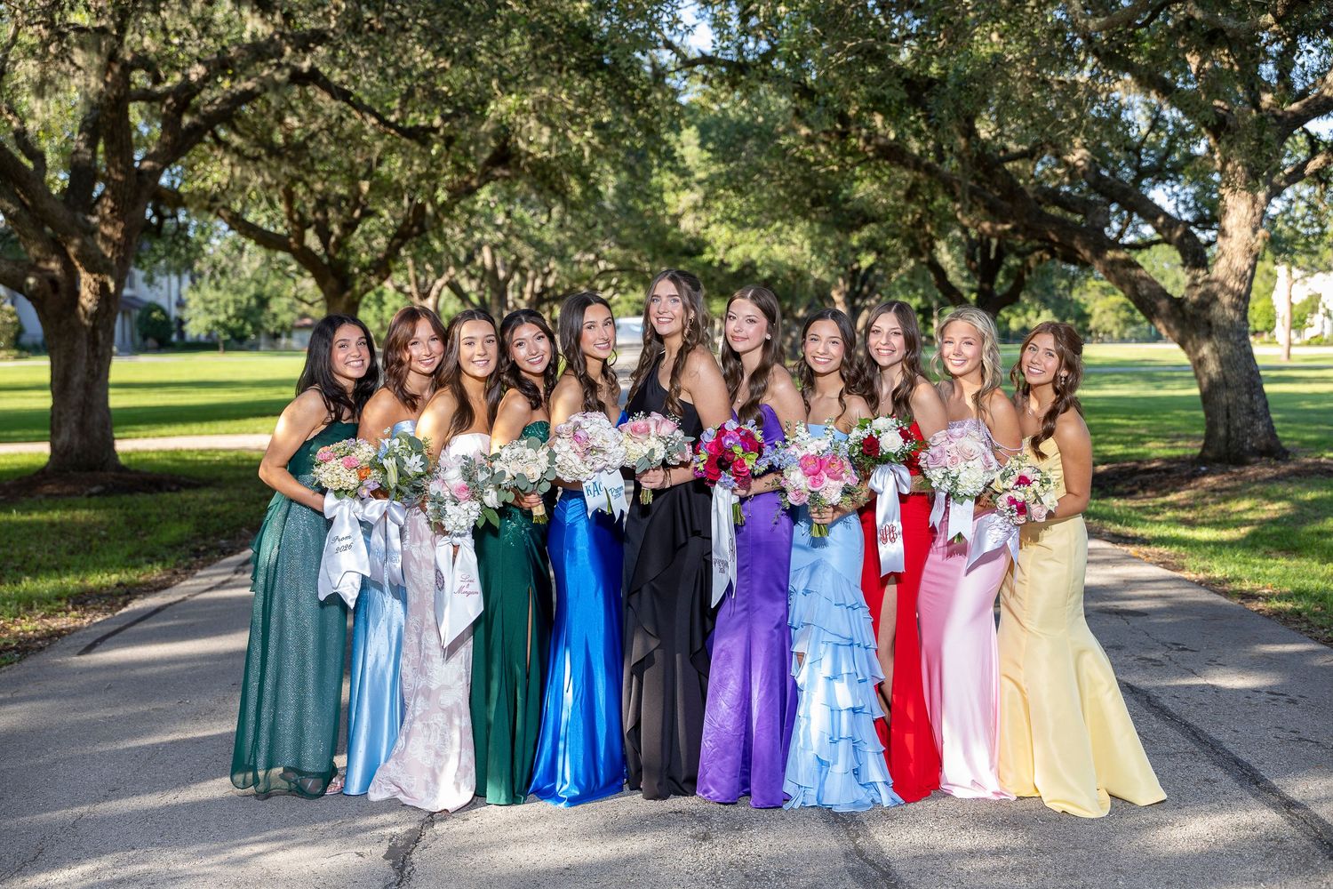 A group of bridesmaids in colorful mismatched gowns holding bouquets pose outdoors under oak trees.