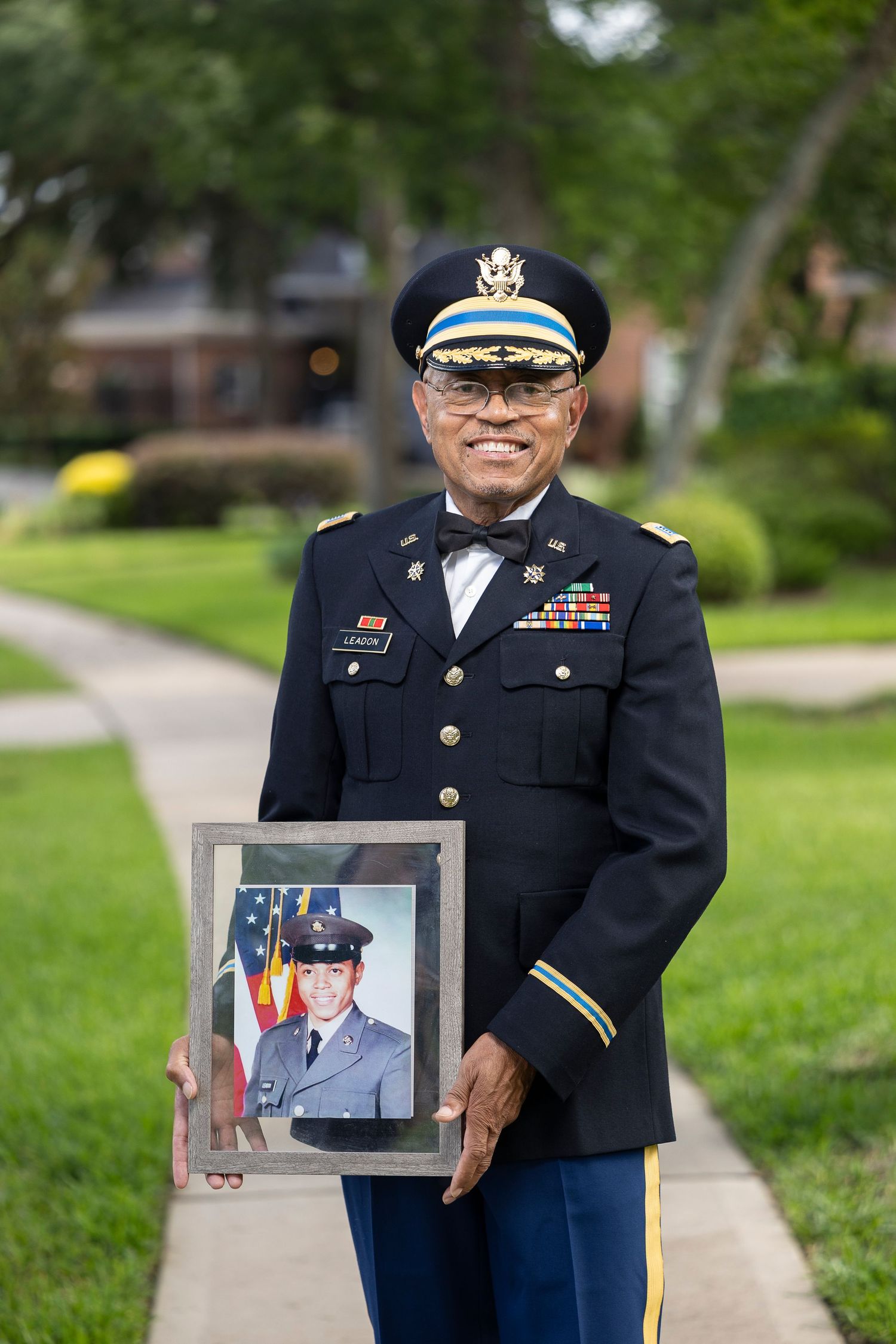 Military veteran in dress uniform proudly holds a framed photo of his younger self in uniform on a suburban sidewalk.