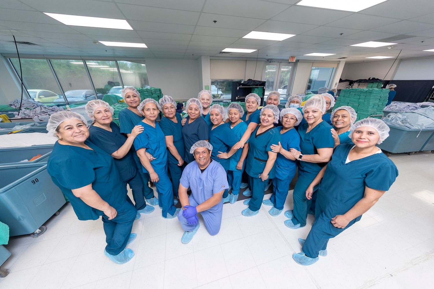 Large group of medical professionals in teal scrubs and hair nets posing together in a modern operating room.