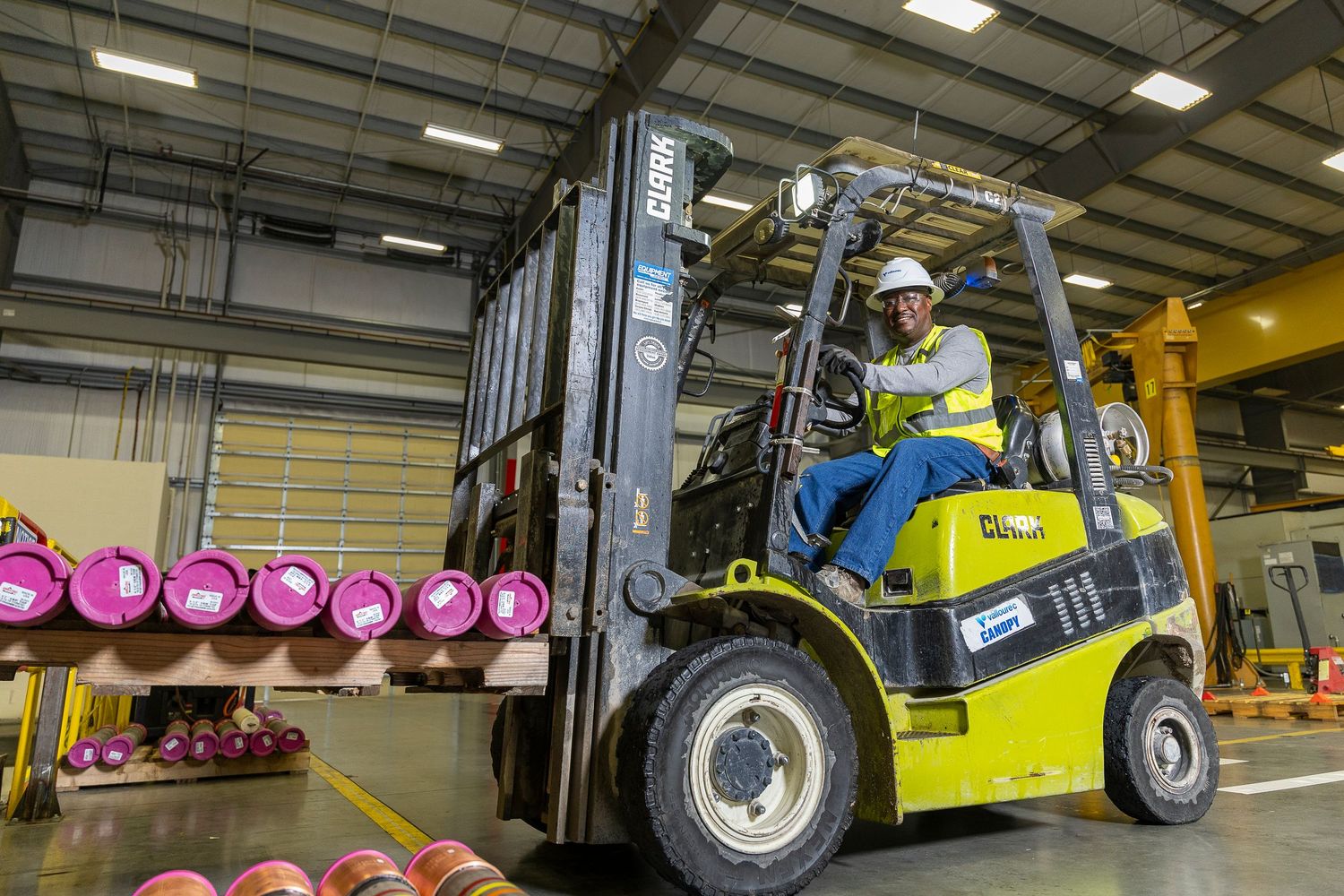 Worker operating a yellow Clark forklift carrying pink rolls in a large industrial warehouse facility.