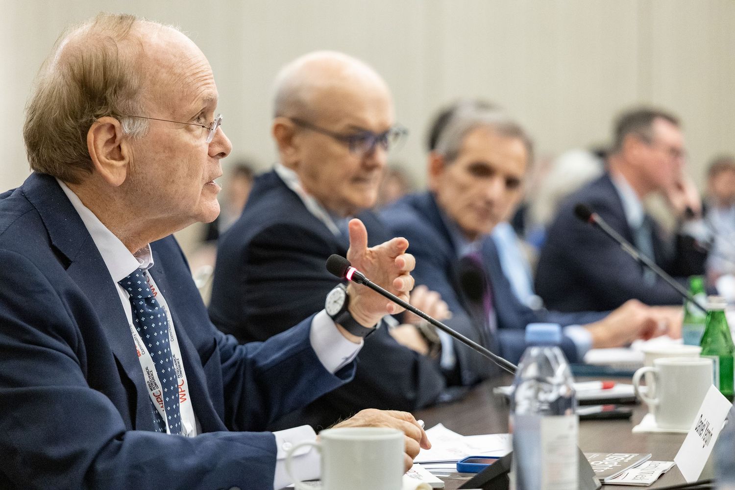 Business professionals seated at a conference table during a formal meeting, with a senior executive speaking into a microphone.