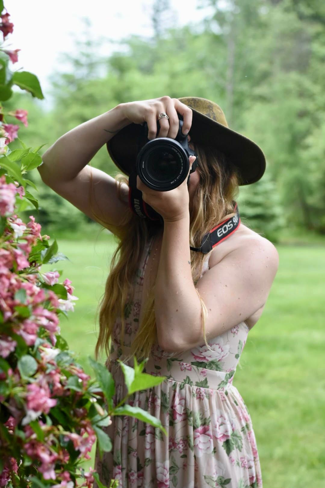 Female photographer in floral dress and hat shooting with a Canon DSLR near pink blooms.