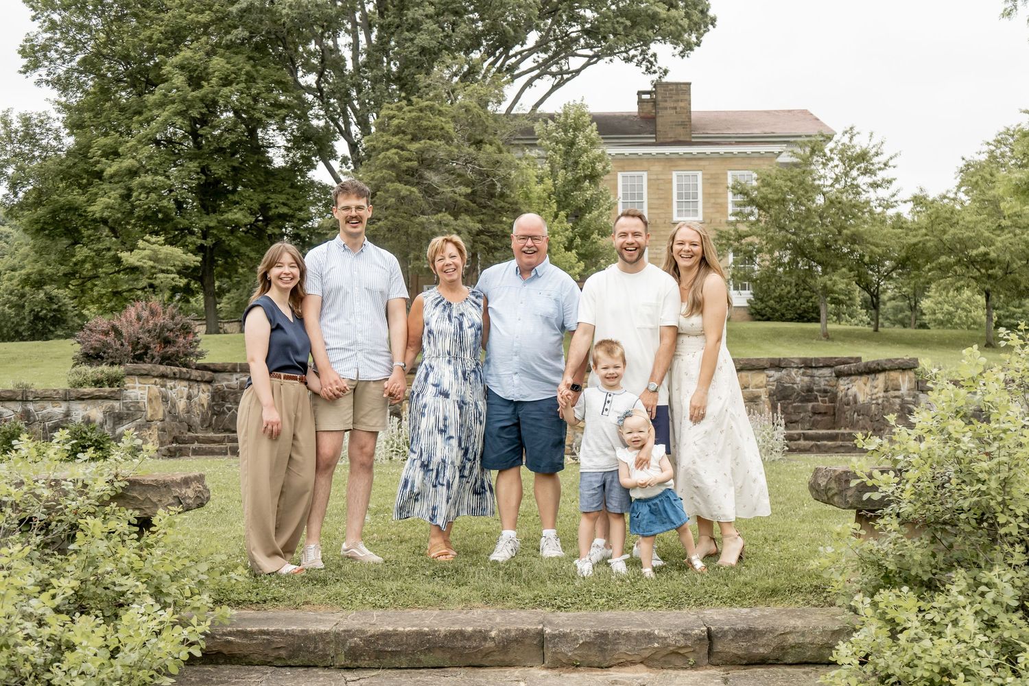Family of seven poses outdoors on stone steps surrounded by lush green trees near a brick building.
