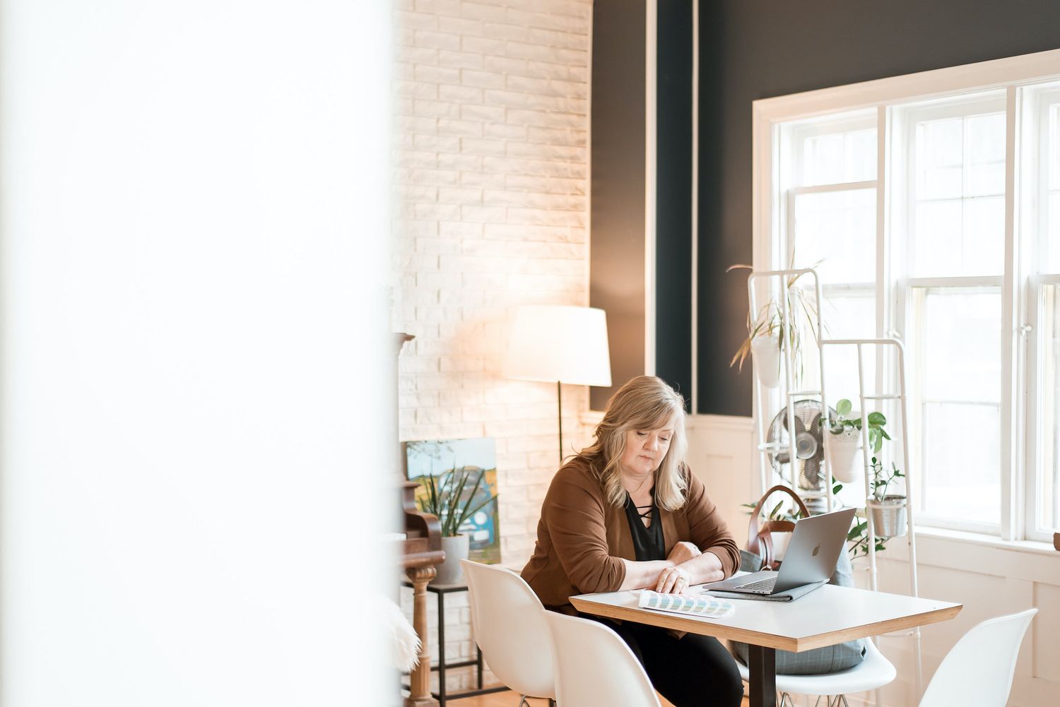 Designer works at modern desk with laptop in bright room with plants and natural light.