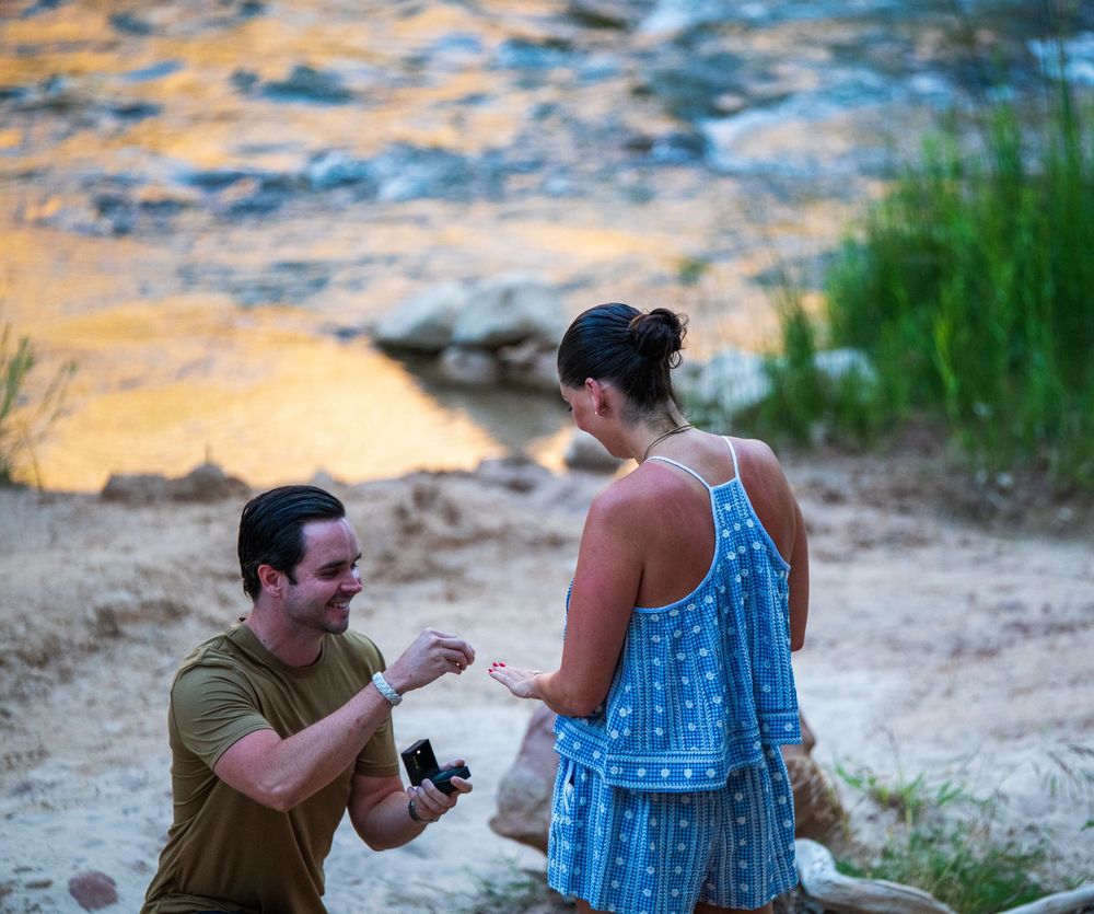 Man kneeling on sandy beach proposing to woman in blue dress at golden sunset by rocky river.