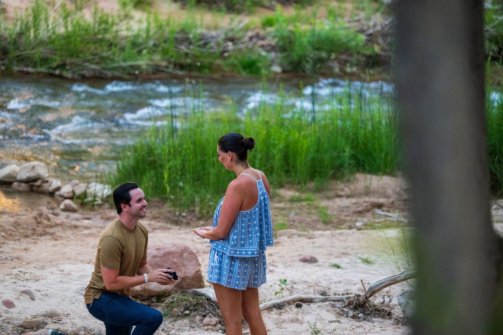 Man proposing to woman by a scenic creek surrounded by lush greenery and sandy banks outdoors.