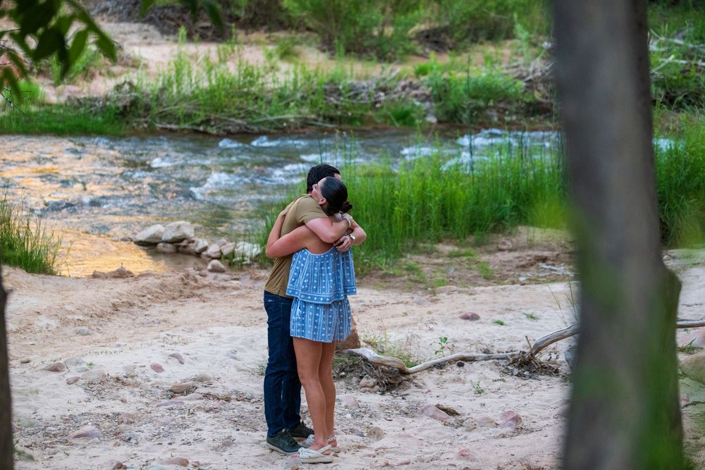 Couple embracing romantically by a serene creek with sandy shores and lush green trees in a natural outdoor setting.