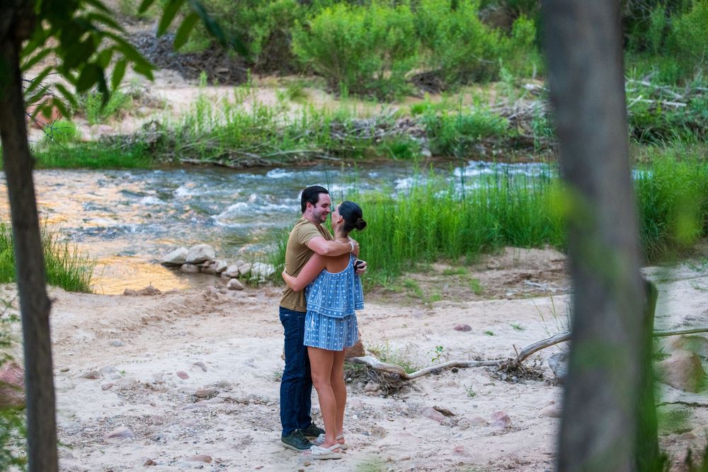 Couple embracing romantically near a serene creek surrounded by lush green trees and sandy banks.