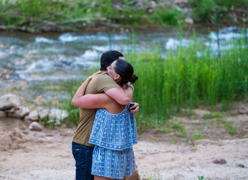 Couple embracing and kissing near a rocky riverbank with lush green grass and flowing water in the background.