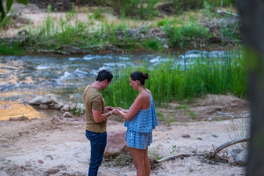 Man proposing to woman by a scenic creek, sharing a romantic moment on sandy riverbank.