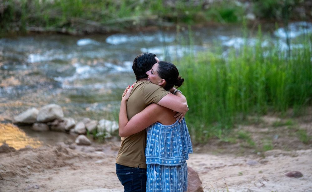 Couple embracing and hugging near a rocky riverbank surrounded by lush green vegetation outdoors.