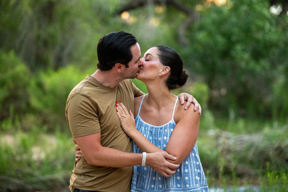 Happy couple sharing a kiss in a lush green forest setting during an engagement celebration.