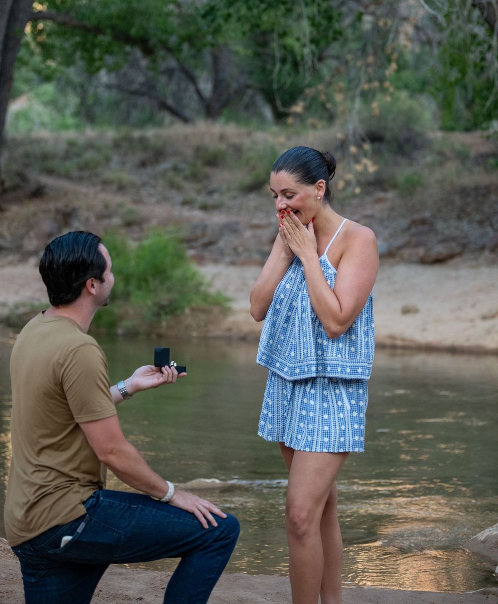 Man on one knee proposing to surprised woman standing in a creek in a wooded outdoor setting.