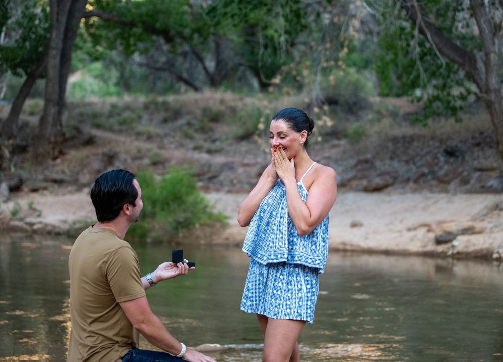 Man kneeling on one knee proposing to a surprised woman by a serene creek surrounded by lush green trees.