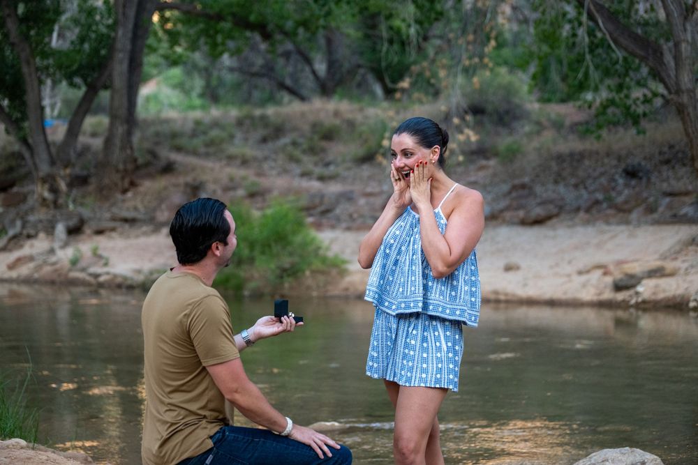 Man kneeling with engagement ring proposing to emotional woman near a rocky creek in nature.