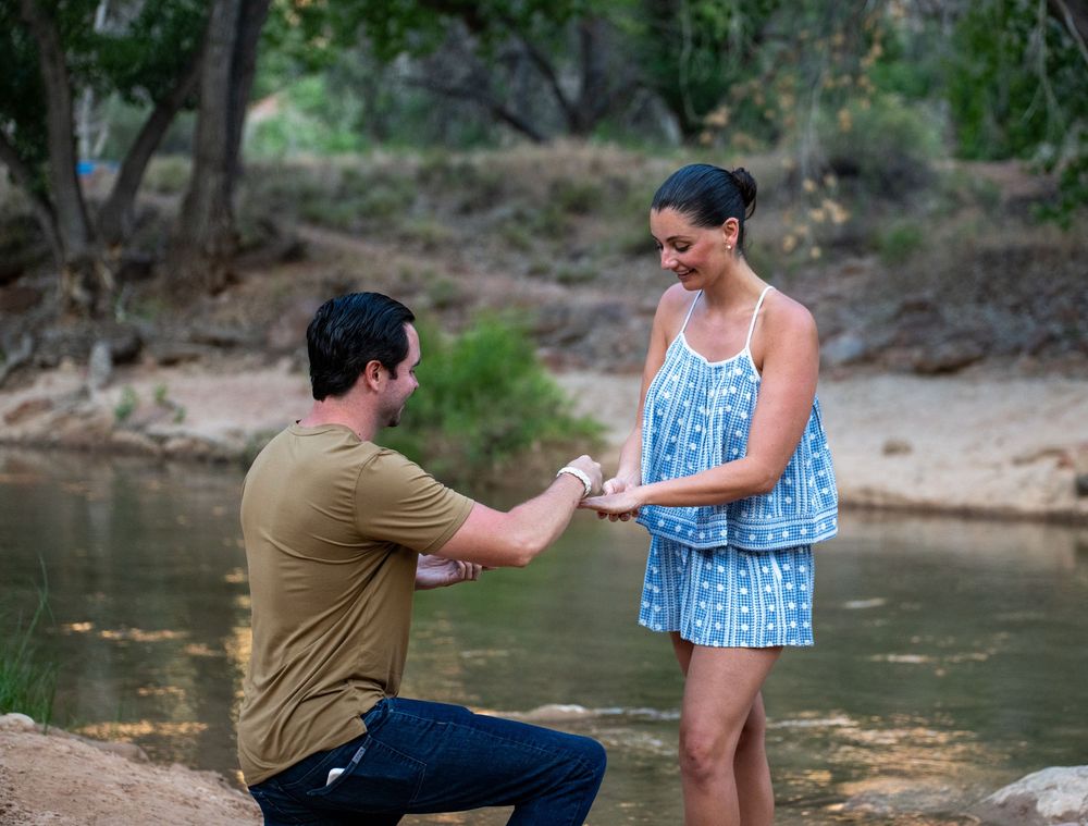 Man proposes to woman on one knee by a scenic river, holding her hands in a romantic outdoor setting.