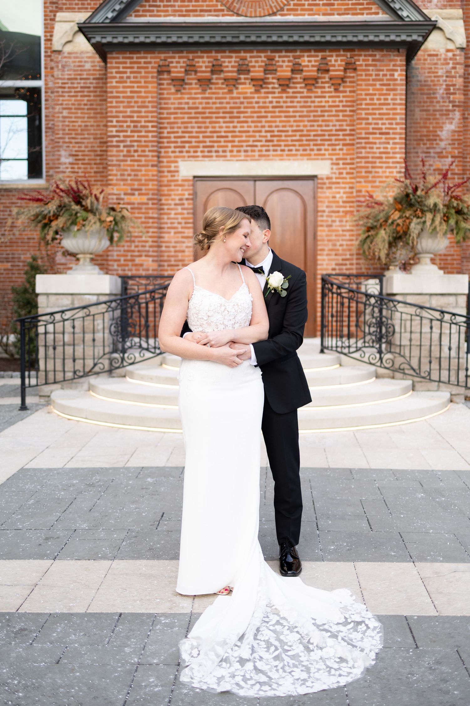 Bride and groom share a romantic embrace outside the Monastery Event Center, bride in lace gown with train, groom in black tuxedo.
