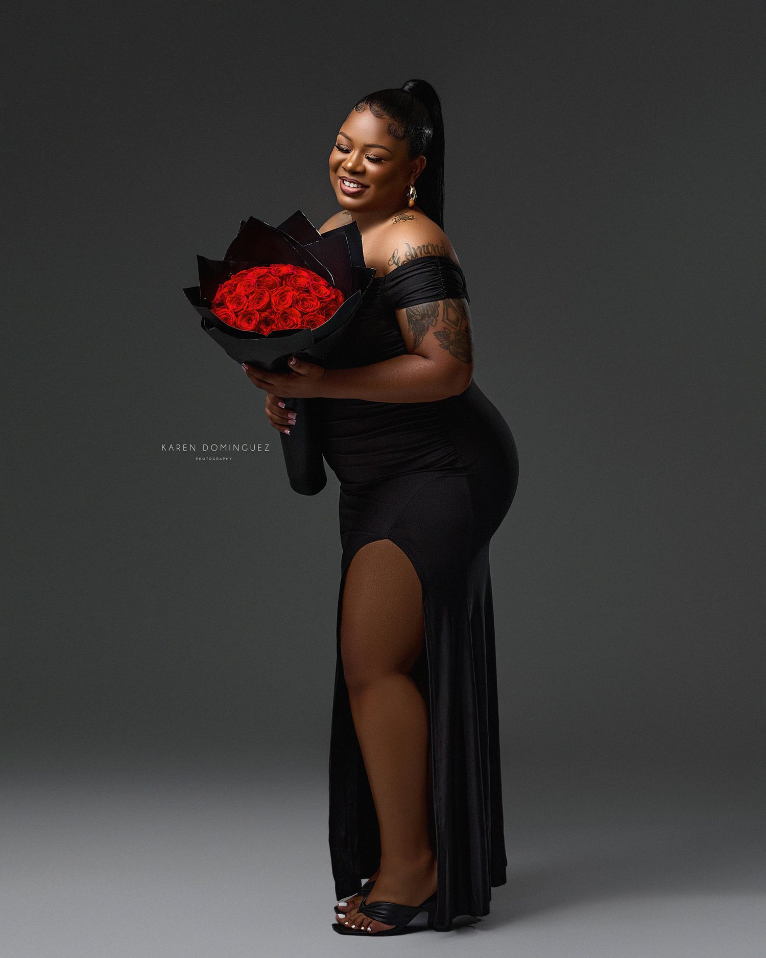 A joyful person in an elegant black dress holds a bouquet of red roses against a dark grey studio background.