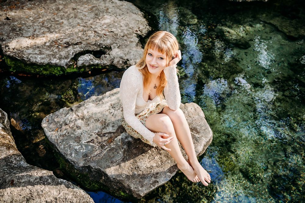 A figure in white sits on limestone rocks beside clear turquoise water in a natural setting.
