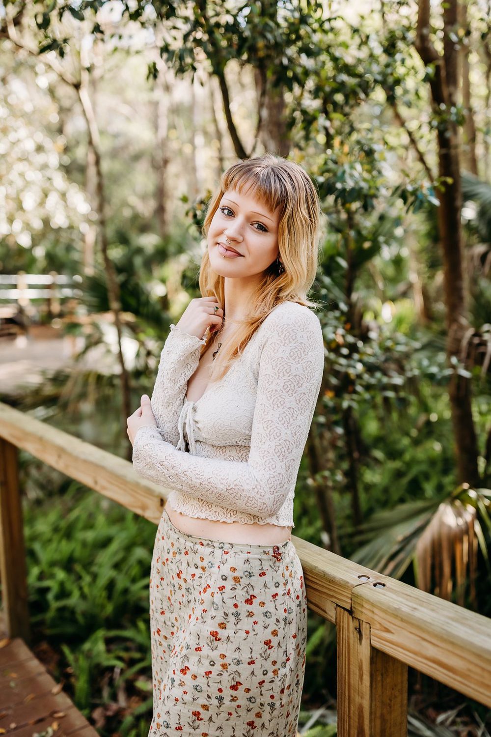 Person in a floral dress poses along a wooden railing in a lush forest setting.