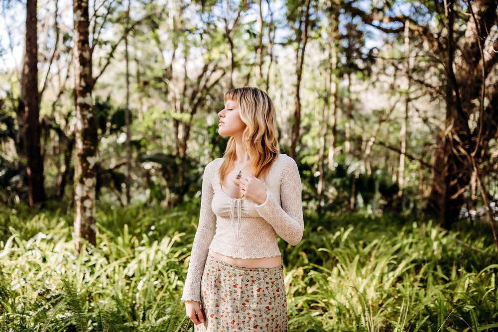 A person stands in a sunlit forest clearing wearing a light-colored sweater and patterned skirt among ferns and trees.