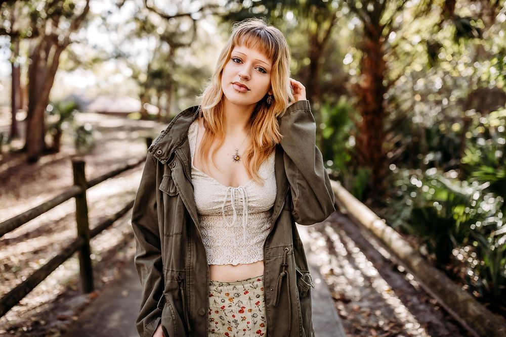 A figure poses in a wooded area wearing a brown jacket and floral dress while sunlight filters through the trees.