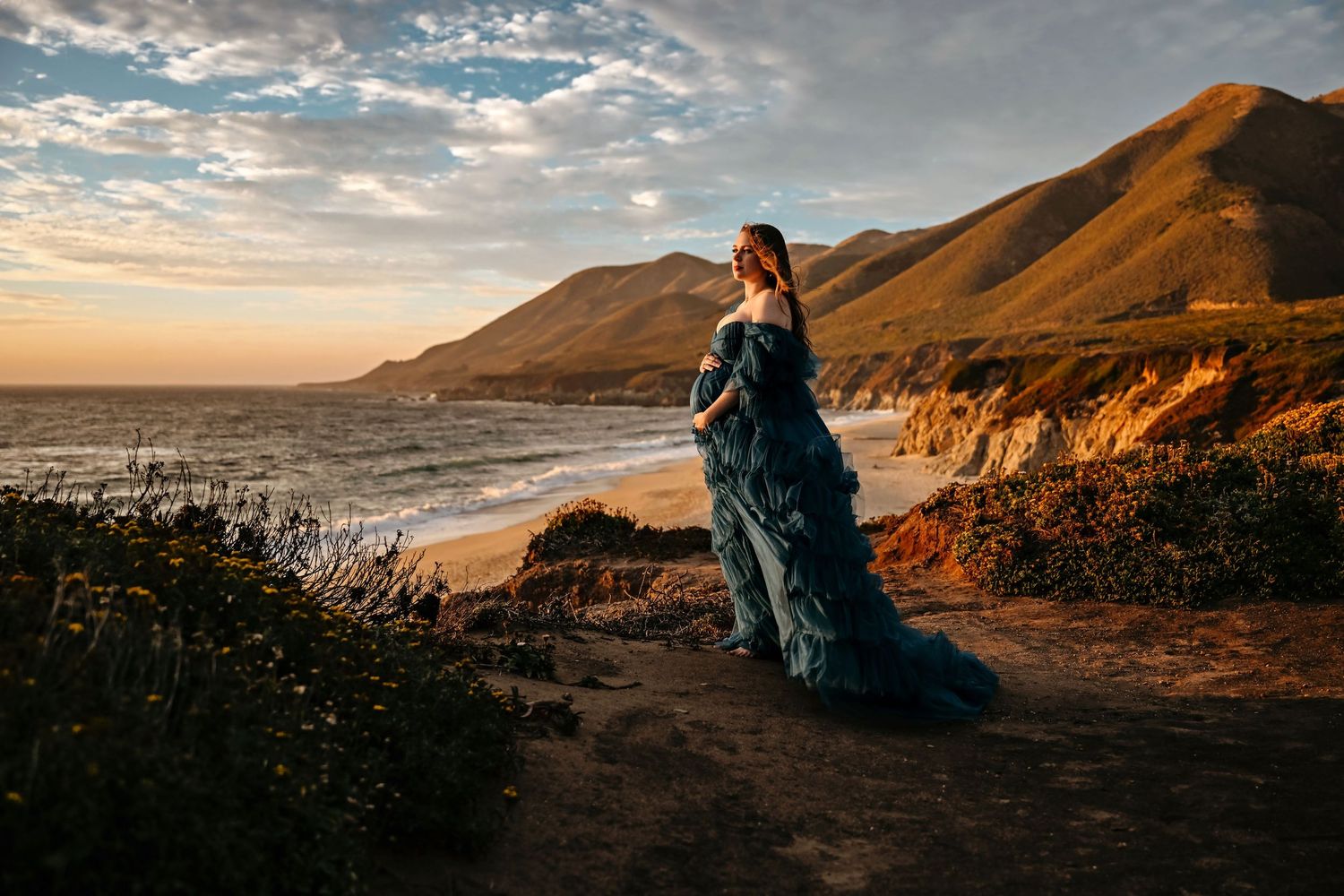 Pregnant woman in teal gown stands on coastal cliff at golden sunset with dramatic mountains and ocean waves in background.
